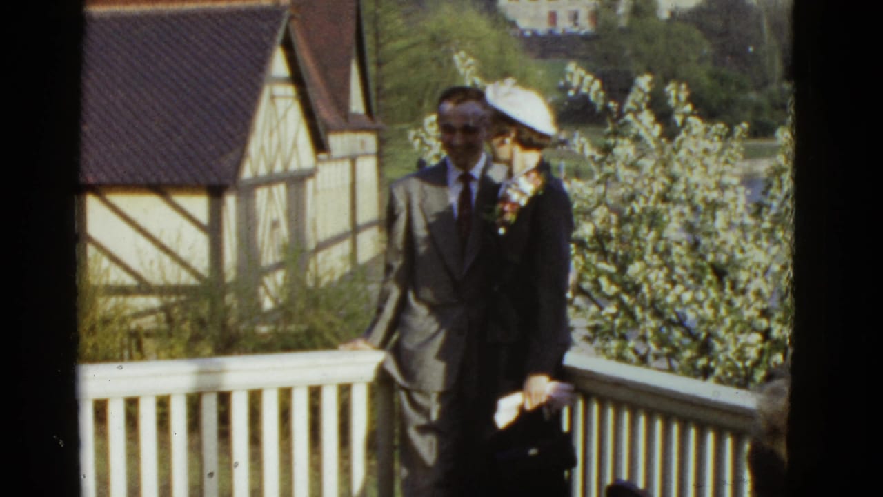 Couple in formal clothing standing by a white balcony railing with trees and houses behind them.