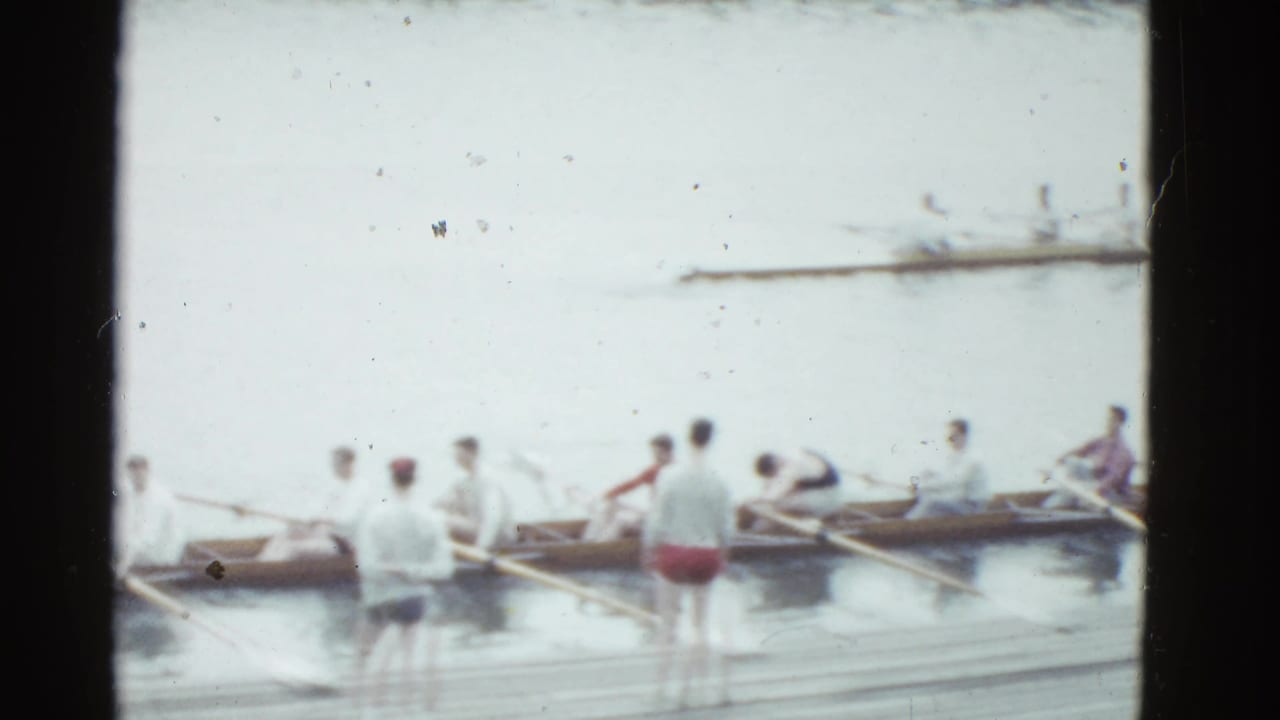 1951 Boston home-movie frame showing rowers in a long shell near a wooden dock, with men standing at the water's edge.