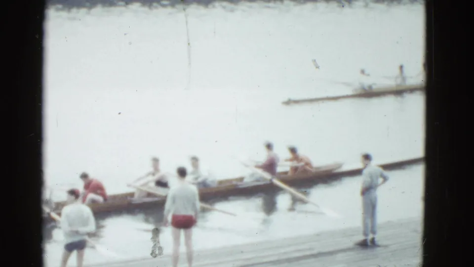 1951 Boston frame showing a rowing shell beside a wooden dock, with several men in light shirts and red or dark shorts watching or preparing.