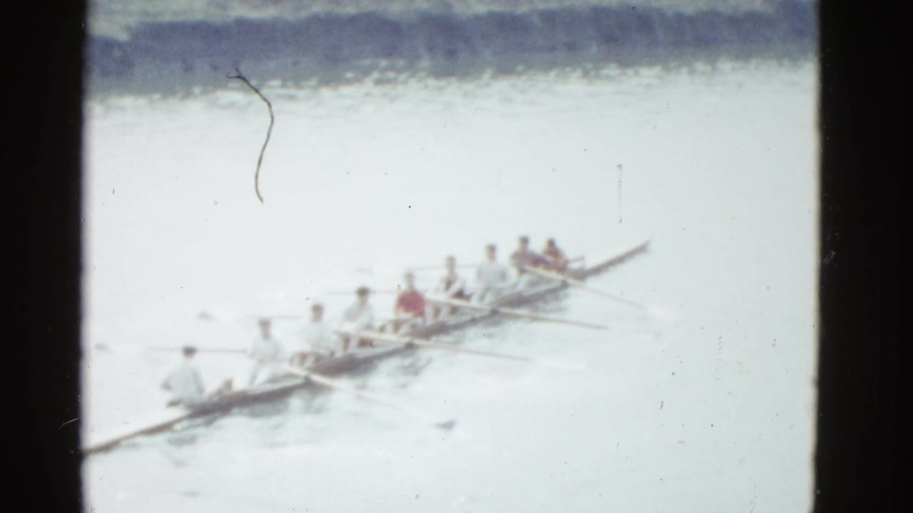 A 1951 Boston-area home-movie frame shows rowers in a long racing shell moving across calm water in soft focus.