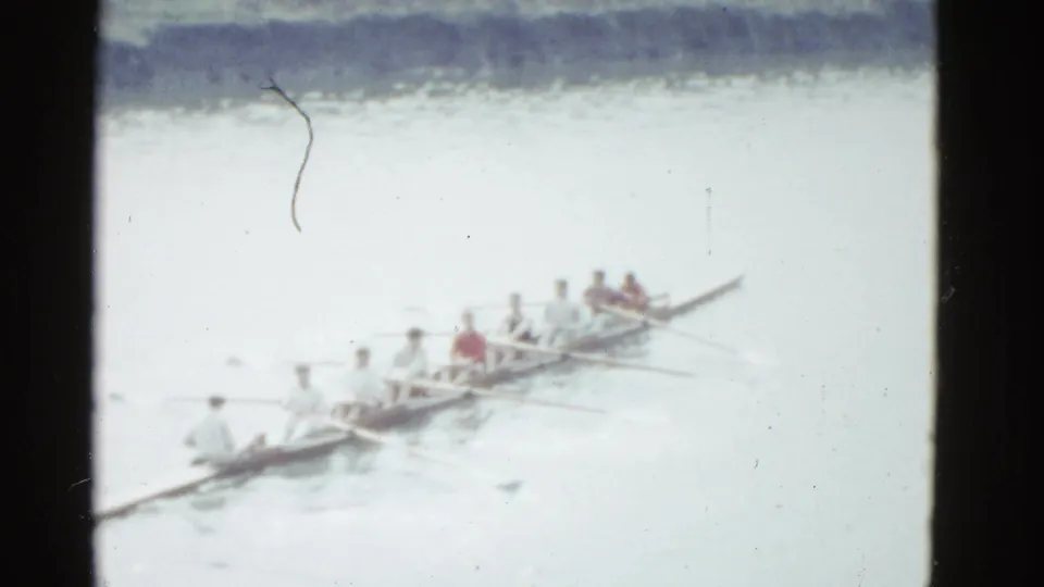 A 1951 Boston-area home-movie frame shows rowers in a long racing shell moving across calm water in soft focus.