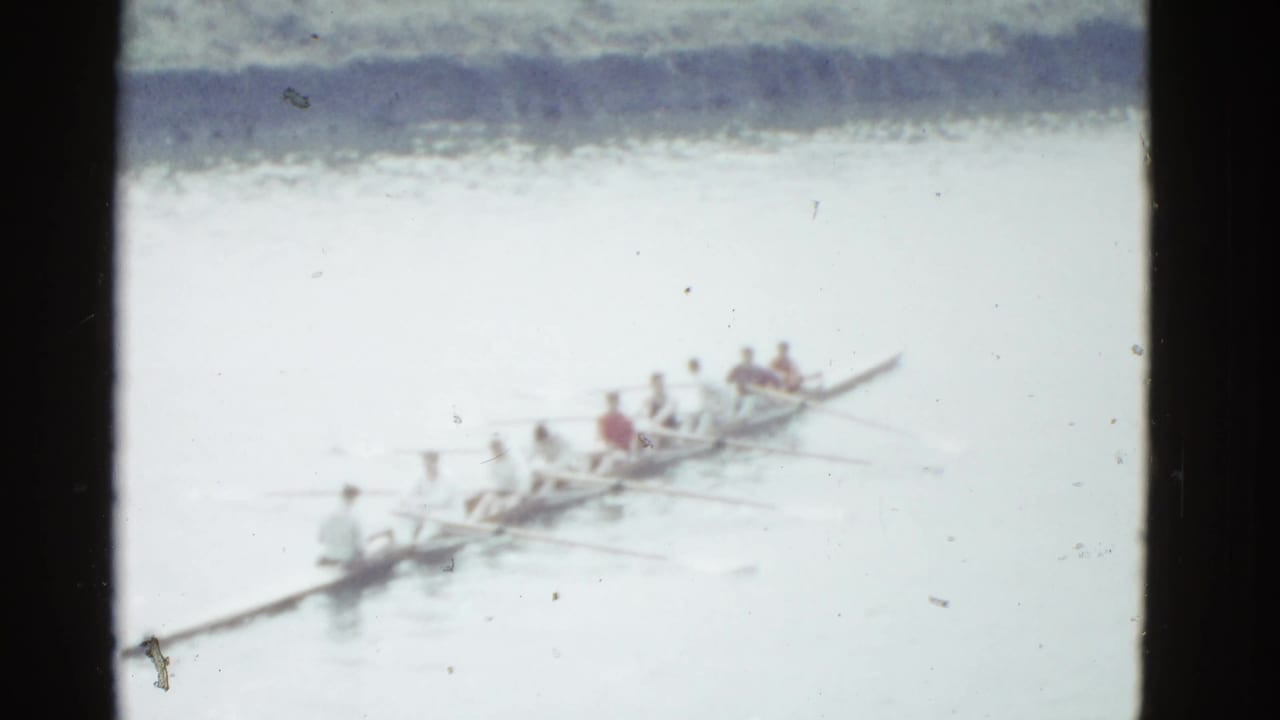 Long rowing shell with several rowers continues across calm water.