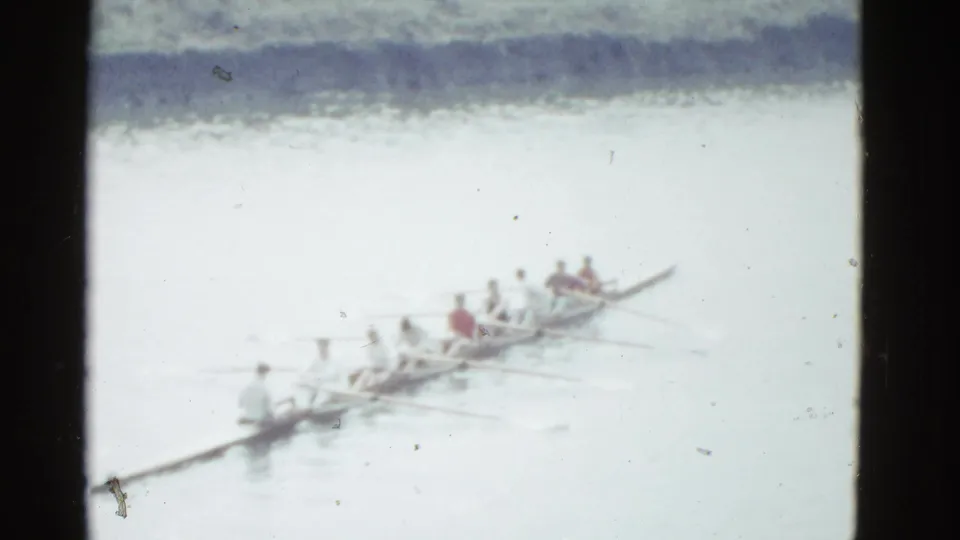 The crew remains visible in a long shell on the water, with oars extended and the shoreline blurred behind them.
