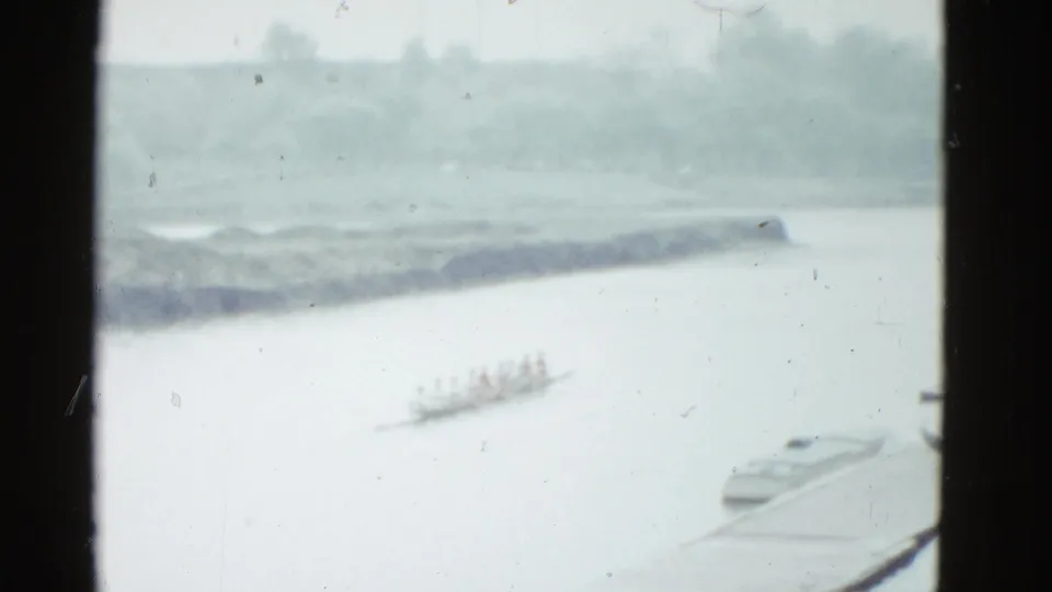 The crew appears farther along the river, partly softened by 8mm grain, with a dark window frame dividing the view.