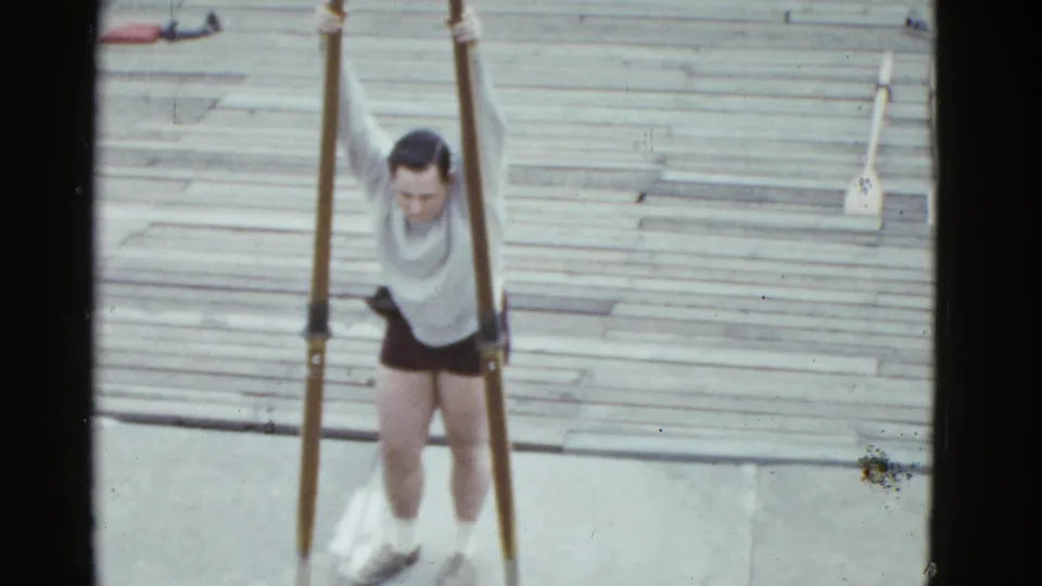 In 1951 Boston home-movie footage, a man in blue shorts stands on a wooden dock holding a pair of long scull oars.