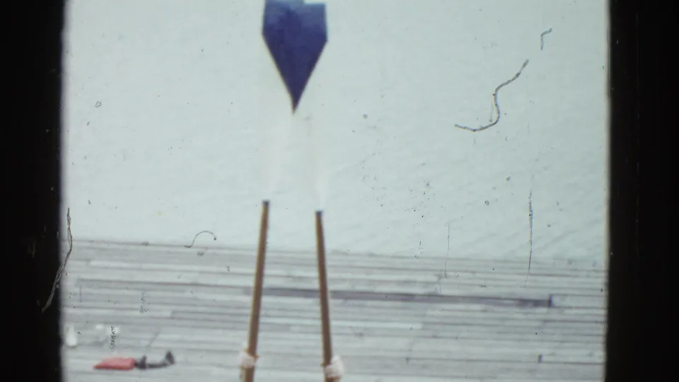 The final frame crops the man low, emphasizing blue shorts, long oars, the dock surface, and pale water in the Boston home movie.