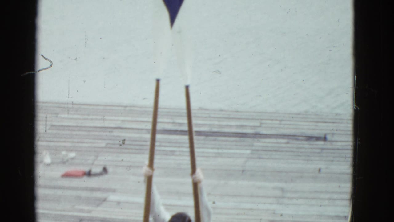 Long oars stand on a wooden pier beside water, with part of a person barely visible at the bottom edge.