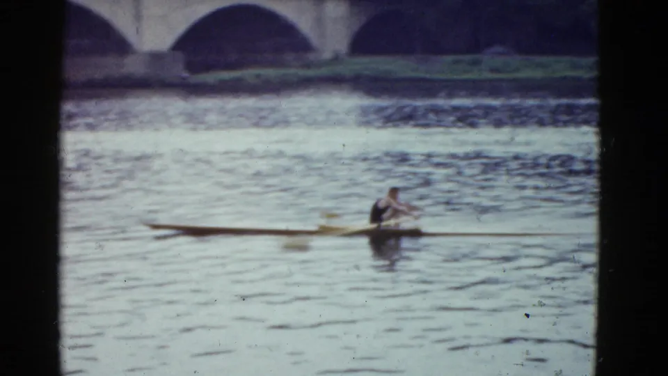 Boston 1951 home-movie frame showing a lone rower in a single scull on the river beneath stone bridge arches.