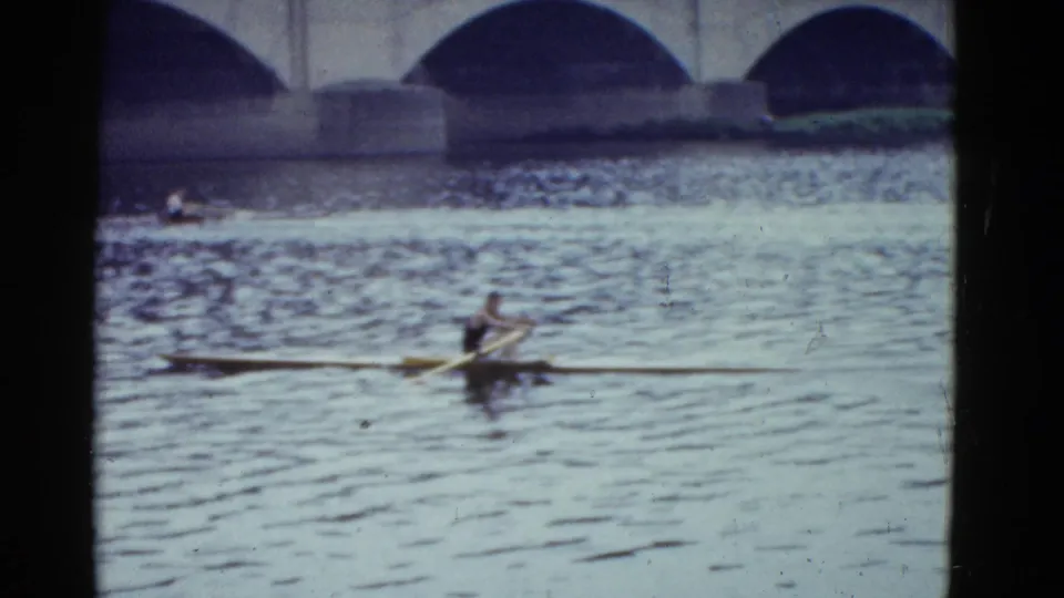 Boston 1951 frame of a rower in a narrow racing shell on the river, with stone bridge arches behind.