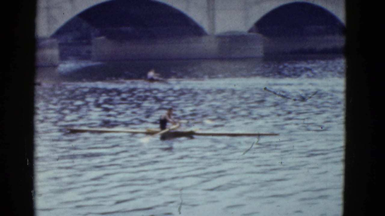 A sculler rows across rippled water beneath a stone arch bridge.