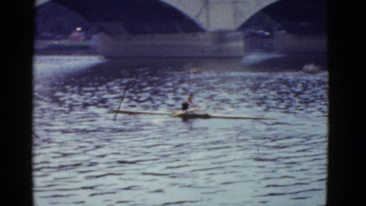 A lone rower glides across Boston river water in a narrow shell with stone bridge arches behind, captured on faded 8mm film.