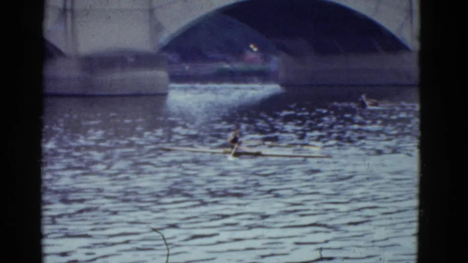 Faded 8mm view of a sculler on a Boston river with bridge arches and distant small craft in the background.