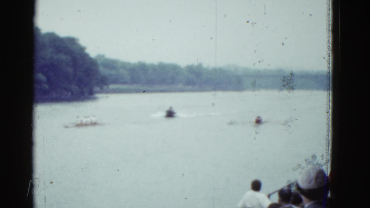 Sculls cross a calm river while people watch from the lower right foreground.