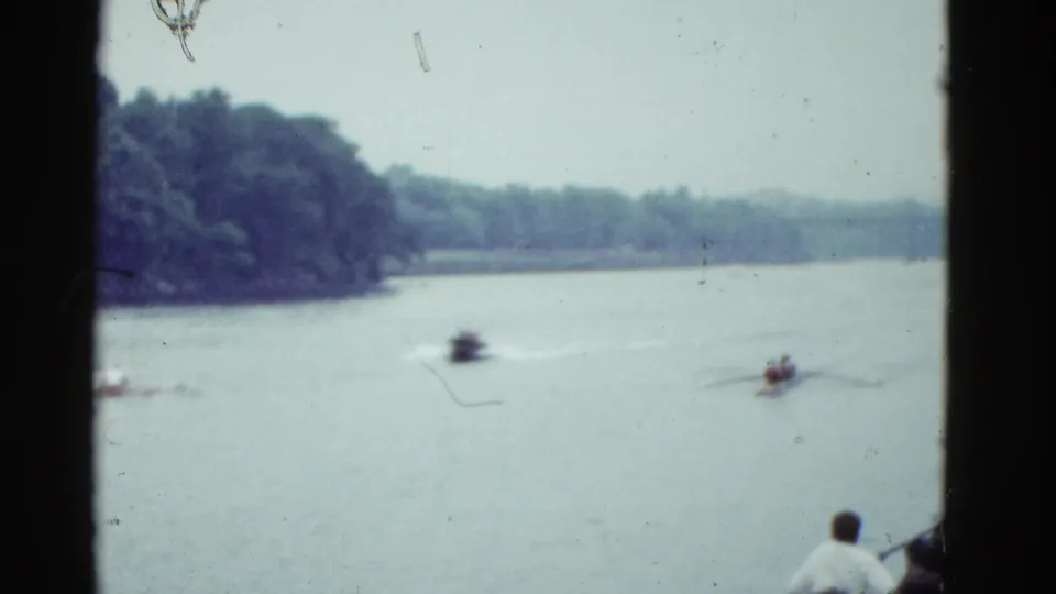 1951 Boston 8mm frame showing a distant boat race on a wide river with wooded banks and spectators partly visible from shore.