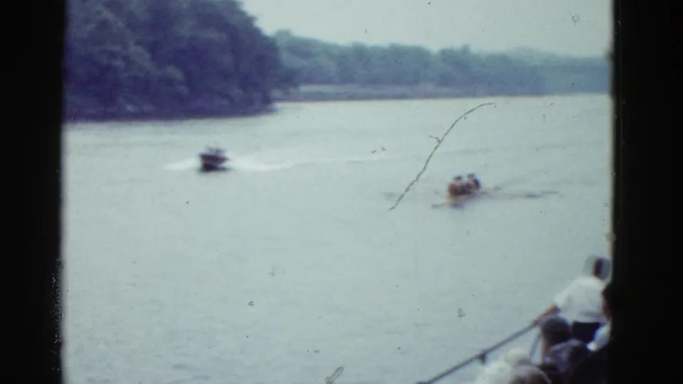 1951 home-movie view of a rowing race from the bank, with a motorboat, crew shell, trees, and foreground spectators.