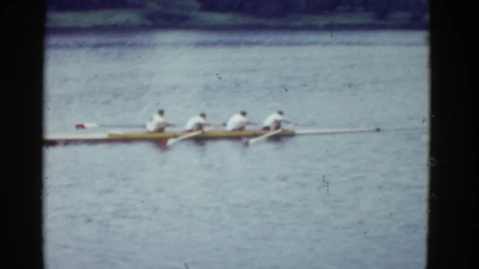 Closer 8mm frame of a four-person rowing shell on a Boston river in 1951, with rowers in pale shirts against rippled water.