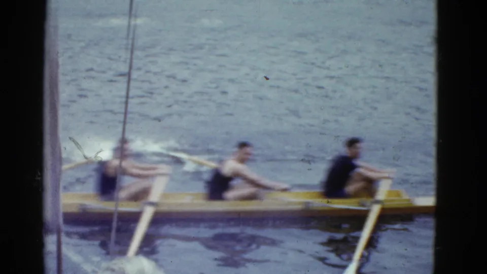 Several rowers sit in a yellow shell, their oars visible as they move across the water in the 1951 Boston clip.