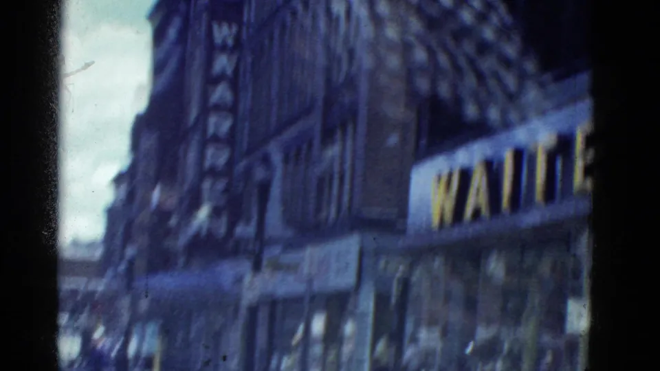 Soft 8mm frame of a Boston commercial street with tall building facades, awnings, traffic, and a partially readable vertical sign.