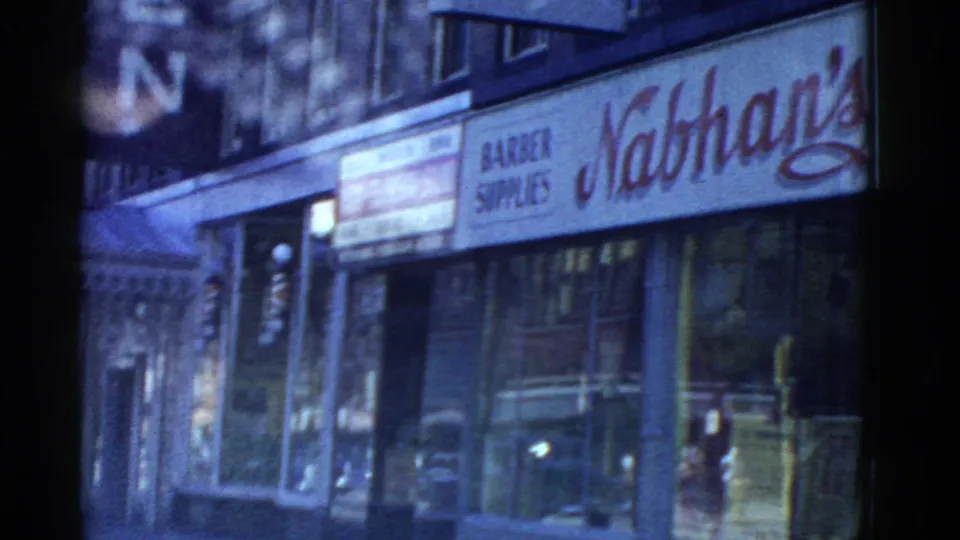 Close street-level 8mm view in Boston showing Nathan's signage, a barber supplies sign, shop windows, reflections, and the lower floors of a commercial building.
