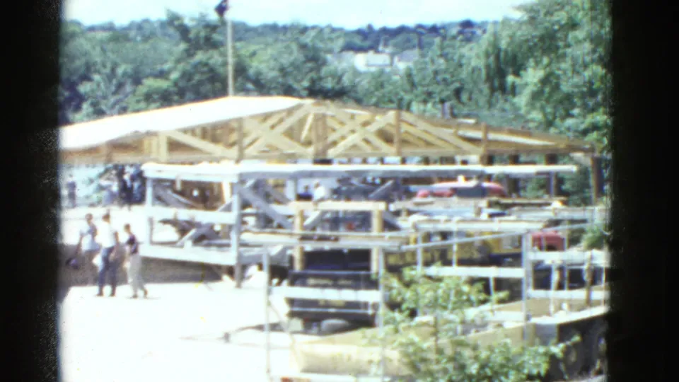 1951 Boston-area 8mm frame showing people near a white-roofed timber-frame construction site with cars and railings in view.