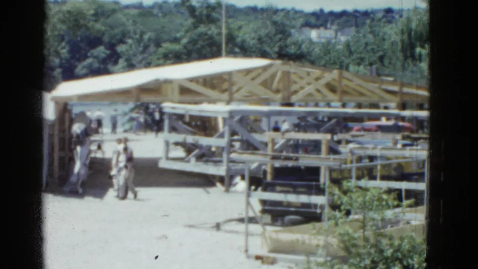 Later frame of the 1951 construction scene, with people in period clothing beside a timber frame and parked cars under bright sun.