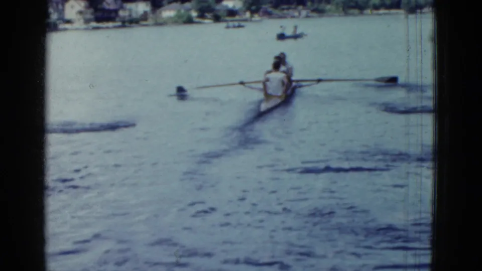 The rowing crew appears farther from view, with shoreline houses or buildings and small boats faintly visible beyond the water.