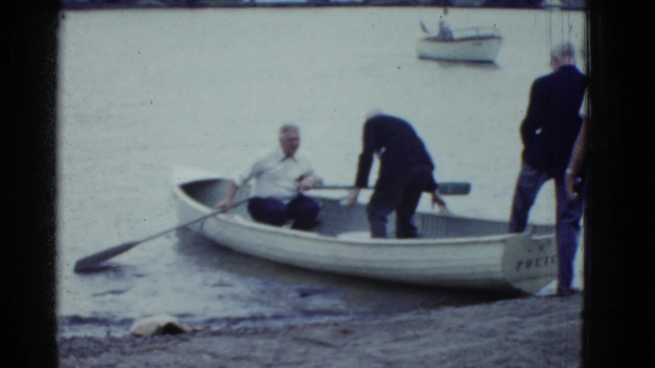 Older men handle a white rowboat at the water's edge while one man sits inside.