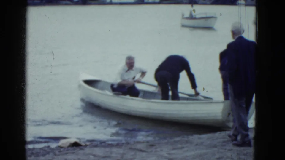 At a sandy shore, older men coordinate around a white rowboat, with one seated at the oars and another bending inside the boat.