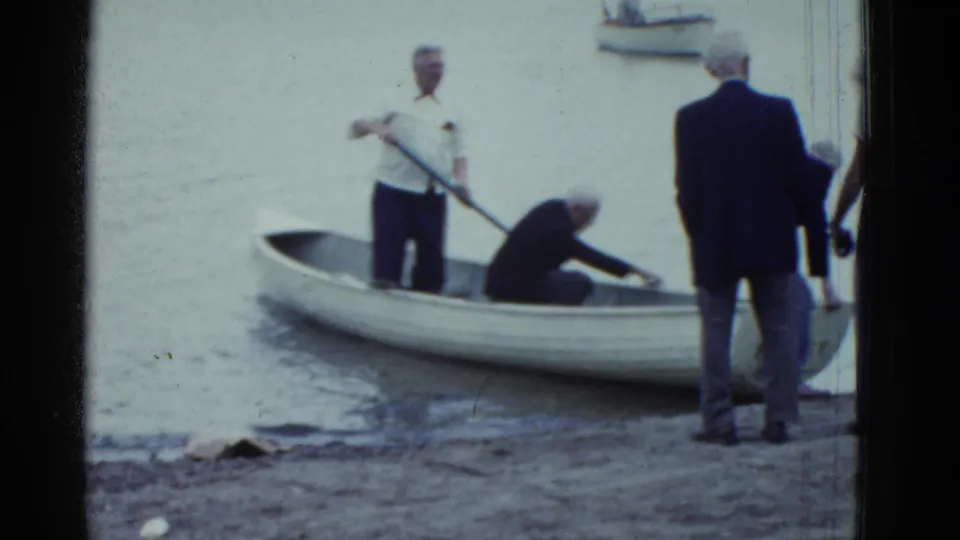 Older men continue preparing a white rowboat at the Boston-area shoreline, one standing with an oar while another reaches inside the boat.