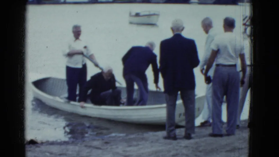 Final 1951 Boston-area frame with men standing and leaning near a white rowboat as a seated passenger adjusts inside the small craft.