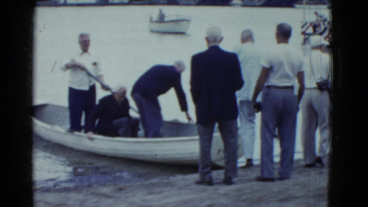 Men gather at a riverbank as an elderly man is helped near a small rowboat.