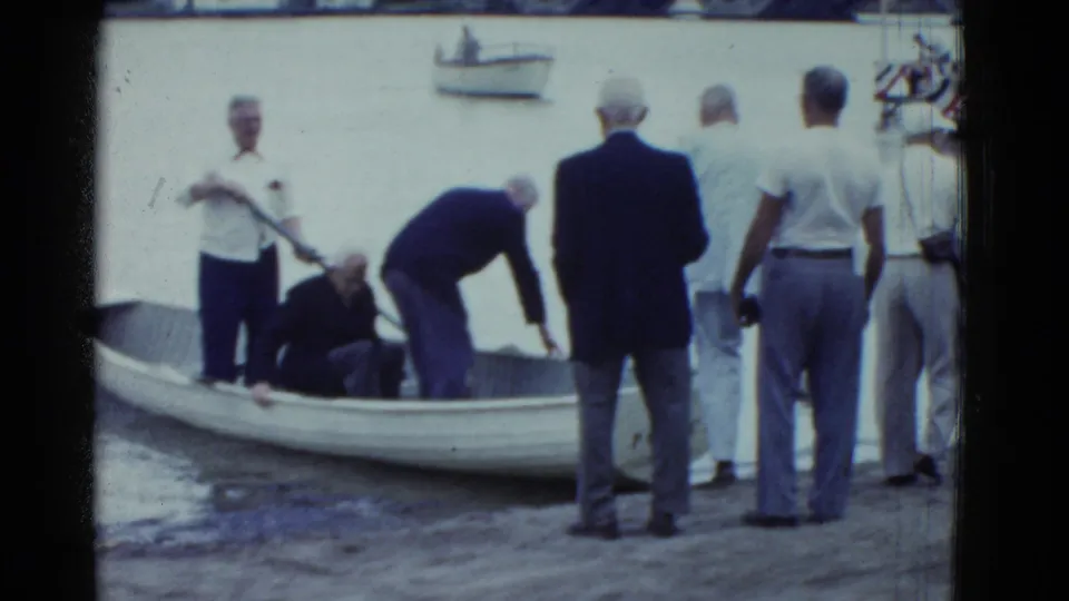 Boston 1951 home-movie frame showing older men at the water’s edge assisting a man beside a small rowboat, with a bridge in the background.