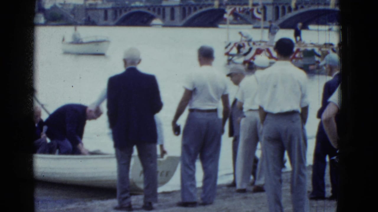 Boston 1951 frame with men in shirts and trousers watching as a man leans into a small rowboat near a decorated waterfront platform.