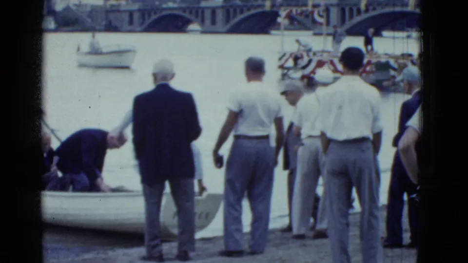 Boston 1951 frame with men in shirts and trousers watching as a man leans into a small rowboat near a decorated waterfront platform.