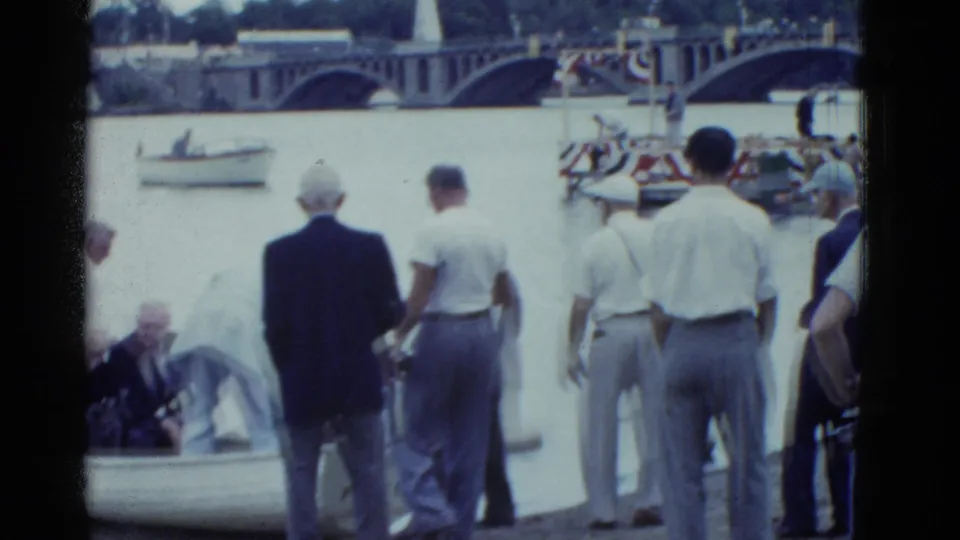 Boston 1951 frame showing a group of men at the river edge, with an elderly man low in the rowboat and a bridge beyond the water.
