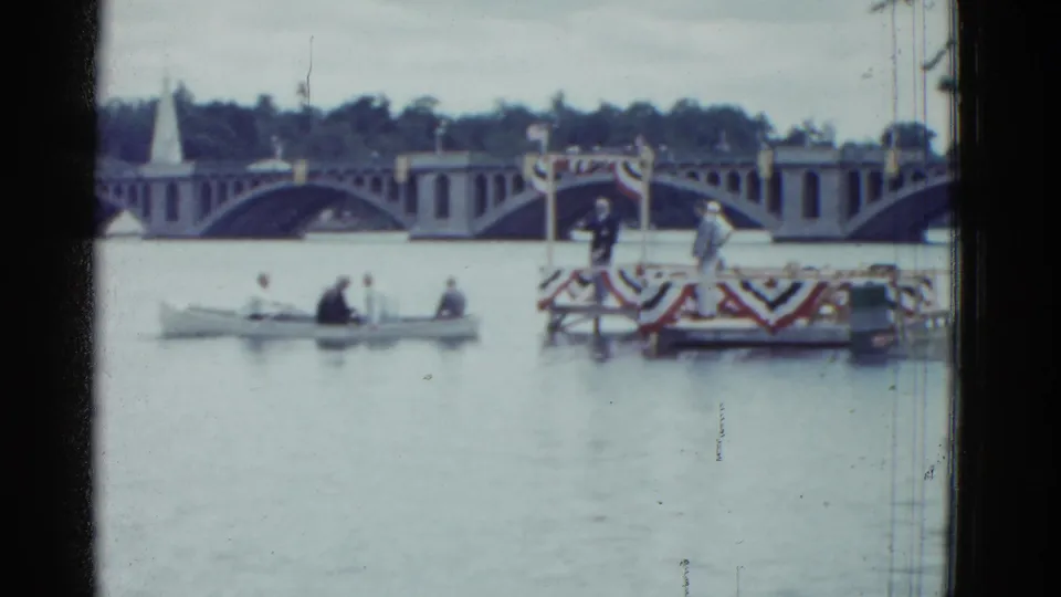 Boston 1951 home-movie frame showing a small rowboat on the water near patriotic bunting and an arched bridge in the background.