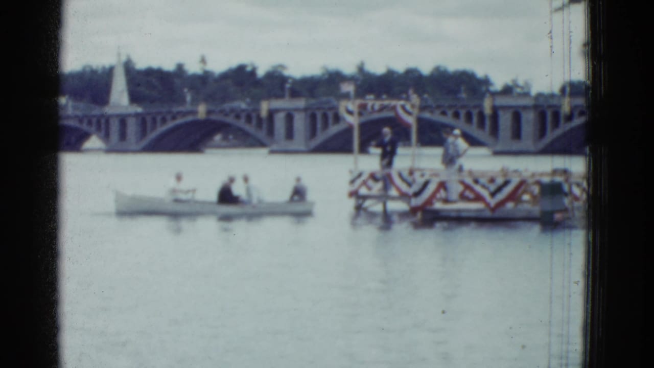 Small boat on a river near a bunting-draped platform and arched bridge.