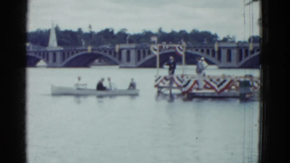1951 Boston home-movie frame showing a small boat moving toward a decorated waterfront platform with an arched bridge behind it.