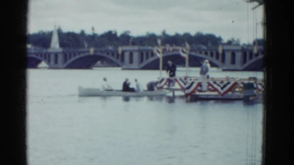 1951 Boston frame showing the boat close to a decorated river platform, with people, bunting, bridge arches, and trees visible.