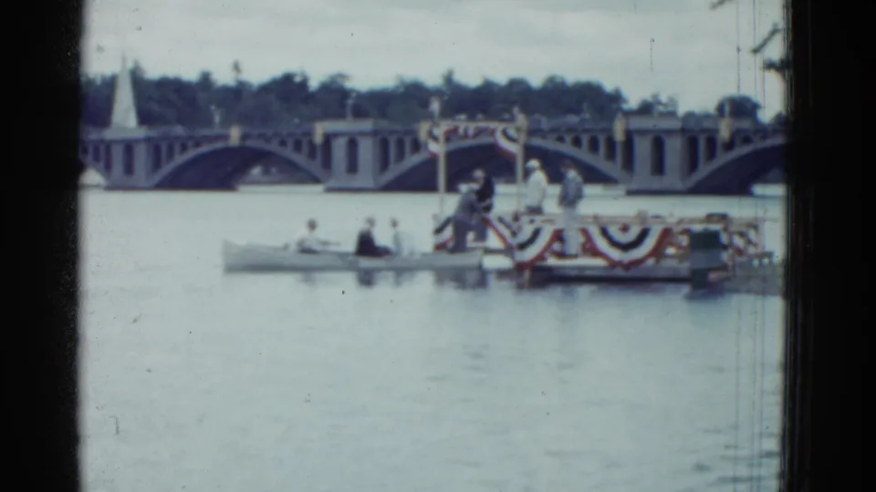 1951 Boston 8mm frame showing a riverside rowing finish area with bunting, small craft, distant people, and bridge arches.
