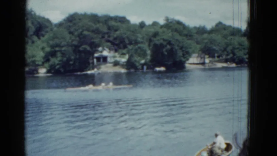 1951 Boston home-movie frame looking across the river toward trees and distant rowing activity, with a small boat cropped at lower right.