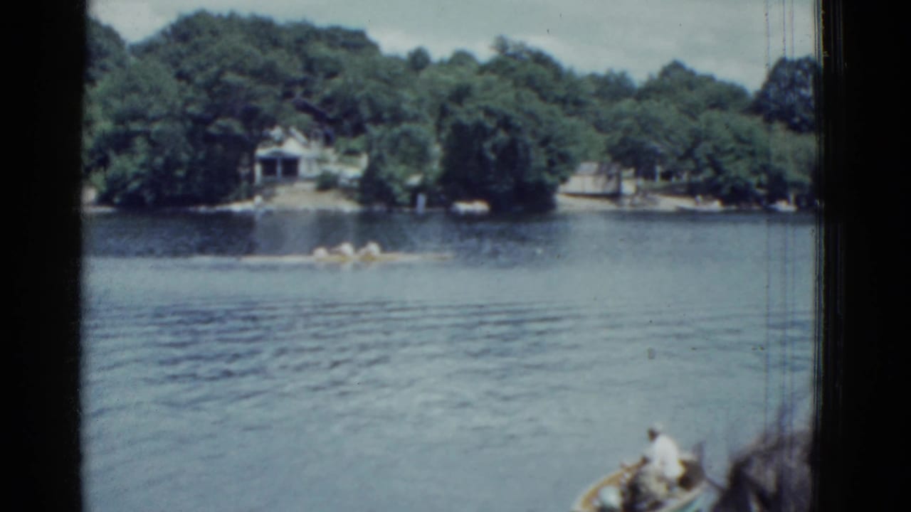 1951 Boston-area 8mm frame showing rowers on a lake, wooded shoreline, houses, and part of a nearby boat in the foreground.
