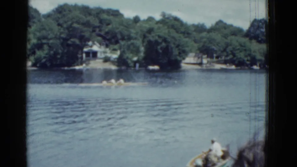 1951 Boston-area 8mm frame showing rowers on a lake, wooded shoreline, houses, and part of a nearby boat in the foreground.
