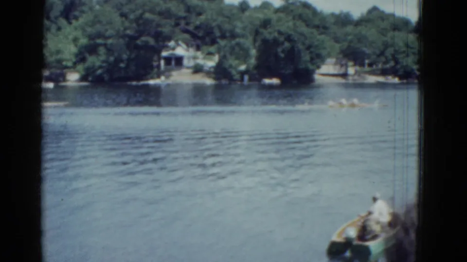 1951 Boston-area 8mm lake scene with a small motorboat near the camera and a long rowboat moving across the tree-lined water.