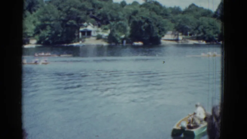 1951 Boston-area lake footage showing a foreground motorboat, distant rowboats, rippled water, trees, and houses along the shore.