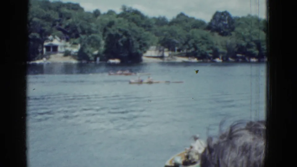 A handheld 8mm view shows distant rowing crews on a Boston lake in 1951, with wooded shoreline and foreground spectators visible.