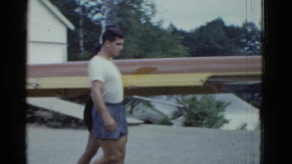 A man in a white shirt and shorts walks past long boats near the Boston lakeshore in 1951 home-movie footage.