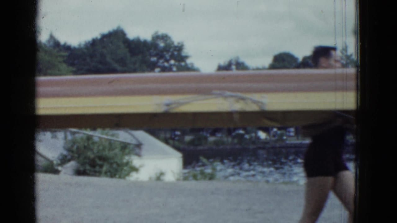 A rowing shell passes across the frame near water with part of a rower visible at right.