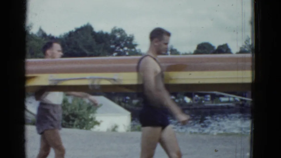Young men in mid-century athletic clothing carry the wooden rowing shell along a waterfront, with trees and water behind them.