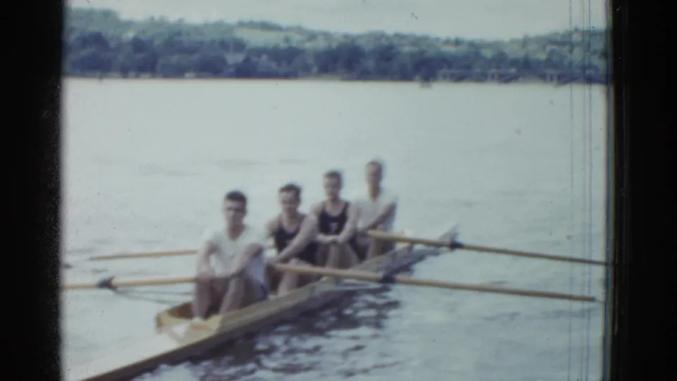 Four young men sit in a rowing shell on the water, holding oars with a wooded shoreline visible in the background.